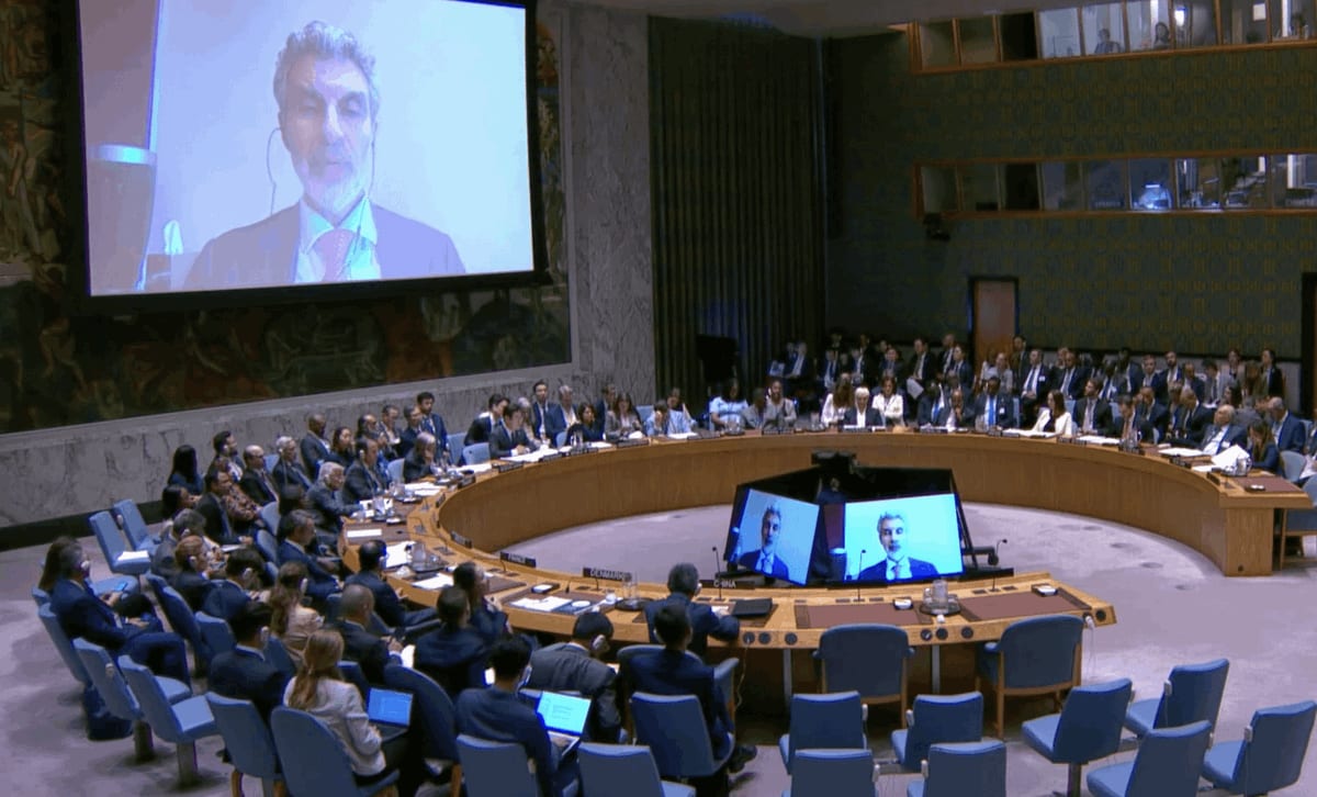 Large room with wooden paneling and large wall screen displaying a man in suit speaking via video. Central round wooden table with multiple screens showing speakers, surrounded by seated delegates in business attire at blue chairs, some with laptops. Additional screens on table display participants. Formal diplomatic setting with flags and microphones visible.